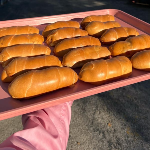 A pink tray filled with rows of golden-brown Venezuelan pan de jamón, held by a person in a pink sleeve.