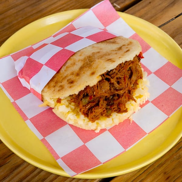 A Venezuelan La Latina arepa filled with shredded beef and cheese, wrapped in checkered paper on a yellow plate.