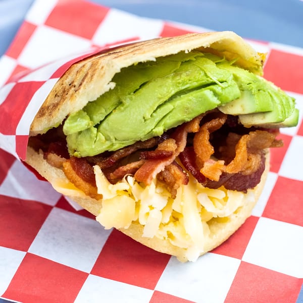 La Latina's Venezuelan arepa filled with avocado, bacon, and shredded cheese, wrapped in checkered paper.